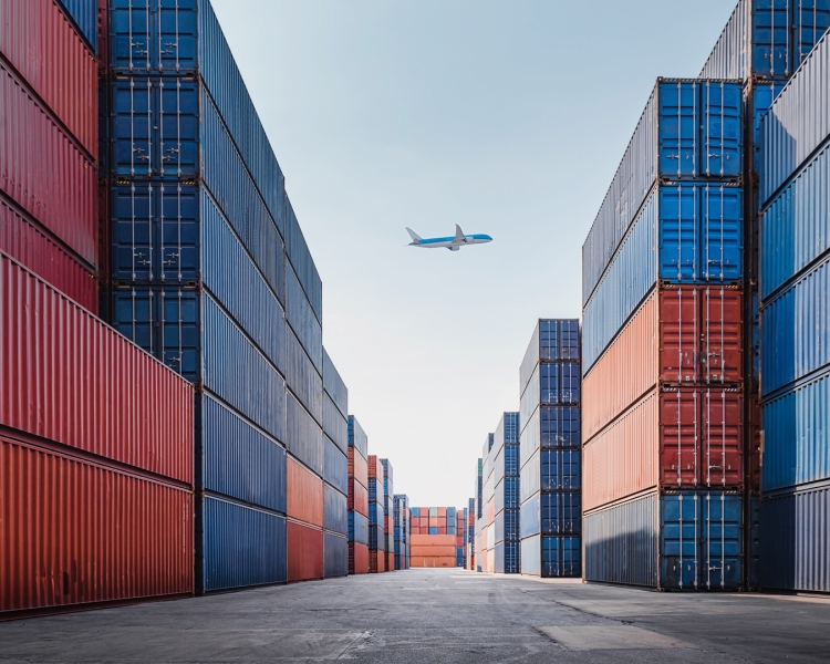 Ein Flugzeug fliegt über Tansport-Container eines Frachthafens