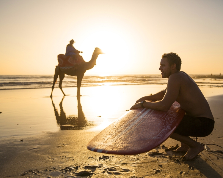 Ein Mann kniet am Strand mit seinem Surfboard in den Händen, dahinter reitet eine Person auf einem Kamel vorbei