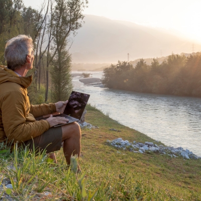Mann sitzt am Fluss mit einem Laptop. Er blickt in die Ferne.