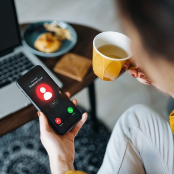 Eine Frau blickt auf ihr Smartphone, da eine unbekannte Nummer anruft und hält eine Tasse mit Kaffee in der anderen Hand