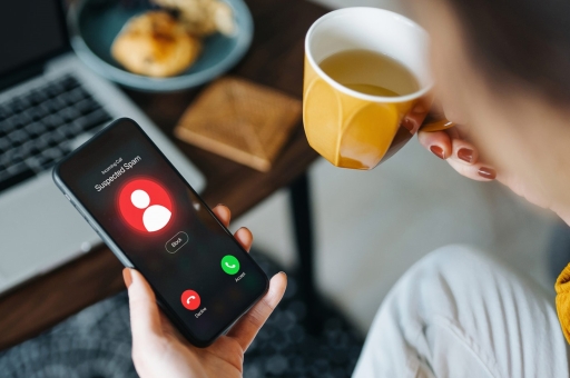 Eine Frau blickt auf ihr Smartphone, da eine unbekannte Nummer anruft und hält eine Tasse mit Kaffee in der anderen Hand