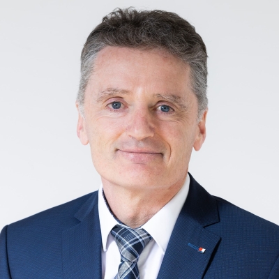 Dr. Hans Eibl trägt einen blauen Anzug, ein weißes Hemd und eine blau gemusterte Krawatte