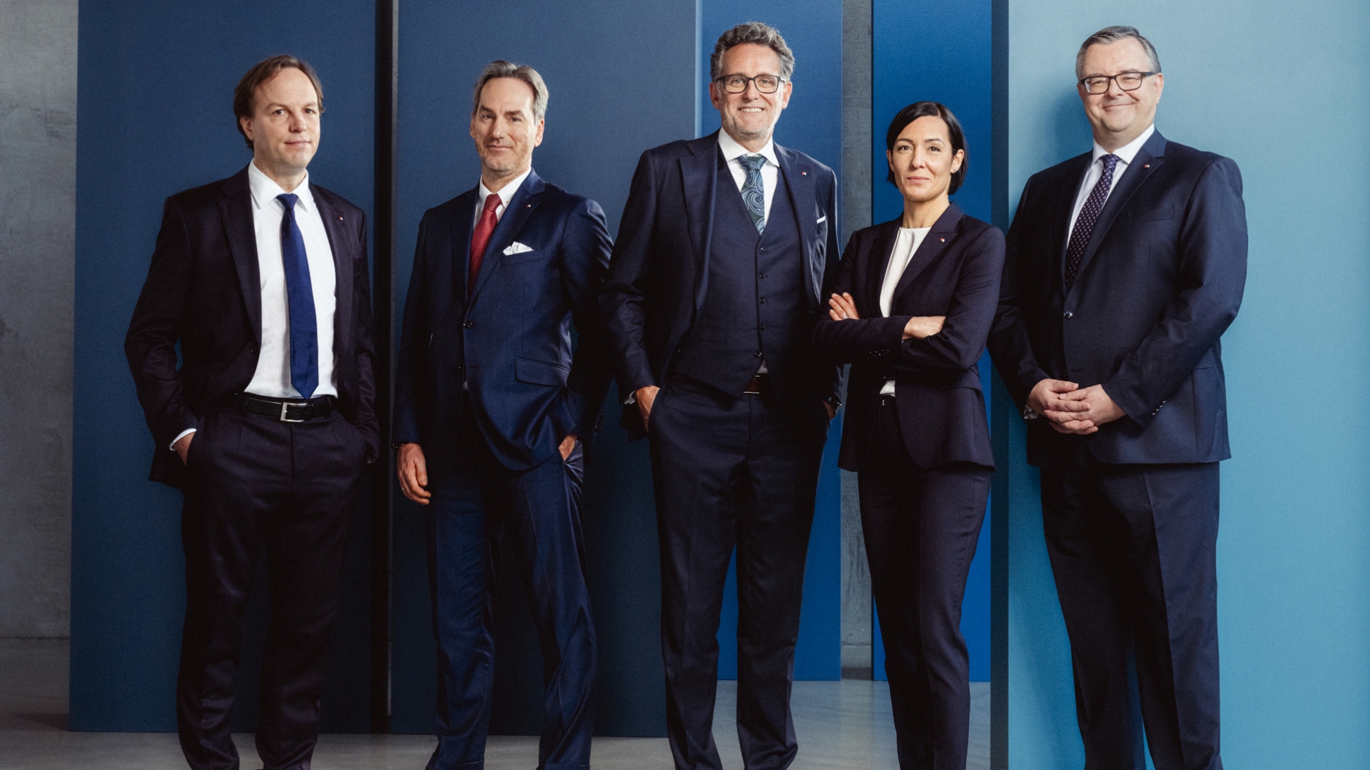 4 Männer und 1 Frau tragen einen Anzug mit einer blauen Wand im Hintergrund.