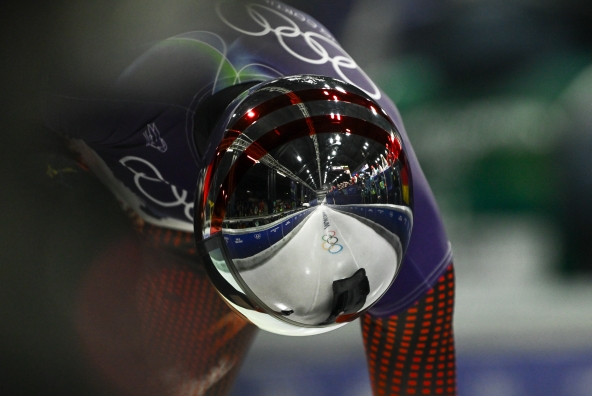 Janine Flock mit Rennhelm und Rennoutfit vorgebeugt in Startposition. Im Helm spiegelt sich der olympische Eiskanal.