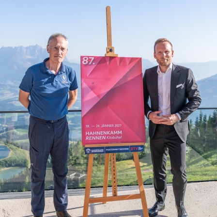 Michael Huber, Präsident des Kitzbüheler Ski Clubs, und Peter Kofler von der BTV präsentieren in Kitzbühel das Hahnenkammposter für die Saison 2027. Auf dem Poster steht: „18.–24. Jänner 2027 – Hahnenkammrennen Kitzbühel“.