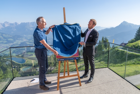 Michael Huber, Präsident des Kitzbüheler Ski Clubs, und Peter Kofler von der BTV präsentieren in Kitzbühel das Hahnenkammposter für die Saison 2027. Auf dem Poster steht: „18.–24. Jänner 2027 – Hahnenkammrennen Kitzbühel“.