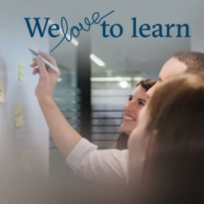Zu sehen sind drei Personen, die auf Post its auf einer weißen Wand schreiben. Darüber steht der Schriftzug We love to learn.